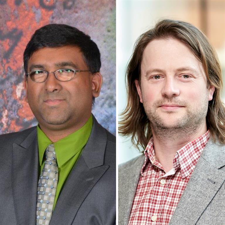 Mahadeo is wearing a black blazer with a green dress shirt and a checkered tie. He is wearing dark glasses, and the background is abstract art. Peter is wearing a grey blazer and a red plaid dress shirt. The background is bright and blurred out. 